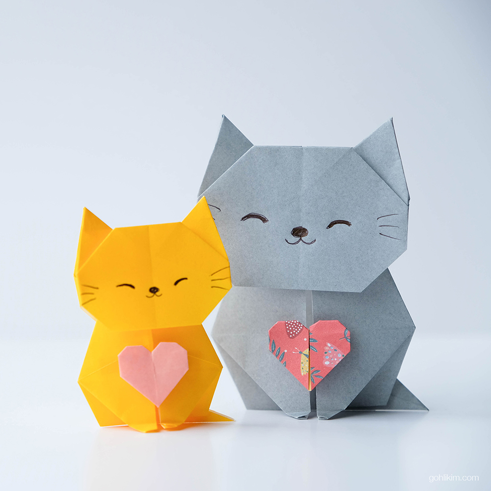 Front-facing origami cat sitting upright with pointy ears, small visible tail, and a tiny paper heart tucked between her delicate front paws.